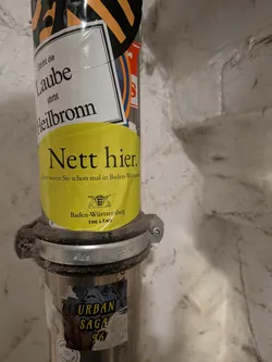 An oval-shaped, bright yellow sticker adhered to a vertical metal pipe. The sticker features the well-known German promotional slogan 'Nett hier. Aber waren Sie schon mal in Baden-Württemberg?' in black serif font, accompanied below by the coat of arms of Baden-Württemberg and the regional marketing campaign text 'Baden-Württemberg THE LÄND'.