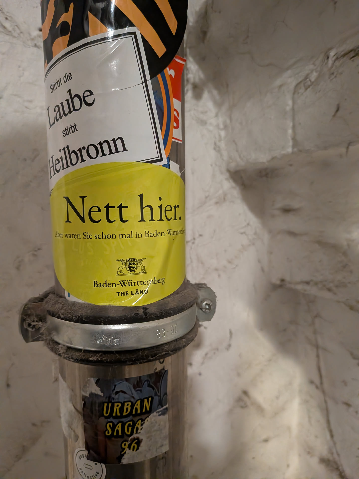 An oval-shaped, bright yellow sticker adhered to a vertical metal pipe. The sticker features the well-known German promotional slogan 'Nett hier. Aber waren Sie schon mal in Baden-Württemberg?' in black serif font, accompanied below by the coat of arms of Baden-Württemberg and the regional marketing campaign text 'Baden-Württemberg THE LÄND'.