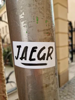 A minimalist sticker with the word JAEGR written in bold, black brush lettering on a white background.  The sticker is affixed to a metal pole.