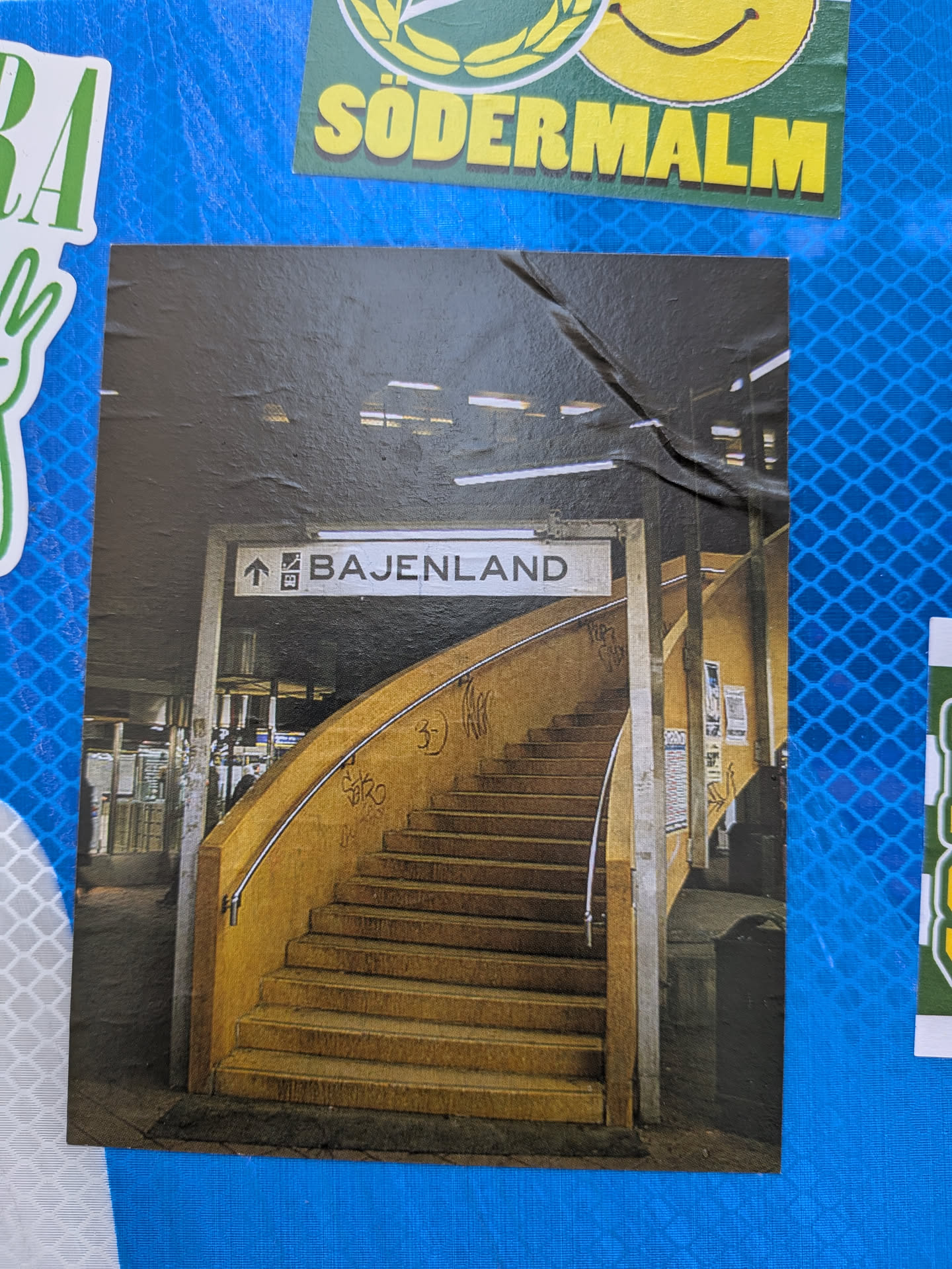 The sticker displays a photograph of a subway or metro station entrance. A prominent, wide, curved staircase with yellow-orange steps and walls dominates the center, leading upwards. A silver handrail runs along the curve of the stairs. Above the staircase, a white rectangular sign with black text and an upward arrow clearly indicates "BAJENLAND". The ceiling is dark with visible fluorescent light fixtures, casting a somewhat dim and industrial light on the scene. The walls surrounding the stairs appear to have some graffiti, adding to a gritty, urban aesthetic. The overall image captures a moment within a bustling public transport environment.