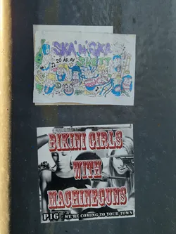 Street sticker The sticker is a black and white image with prominent red text overlay. It depicts two women; the left one is shown in a partial profile, looking downwards, while the right one is shown from the chest up, looking directly at the viewer with a slight smile, with what appears to be a stylized machine gun near her face. The title 'BIKINI GIRLS WITH MACHINEGUNS' is in large, embossed-looking red letters, dominating the top half of the design. Below the women, in smaller white text with a black outline, is 'P1G - WE'RE COMING TO YOUR TOWN', suggesting a band name and a tour or presence. The overall aesthetic hints at a grindhouse movie poster or punk/rock band imagery.