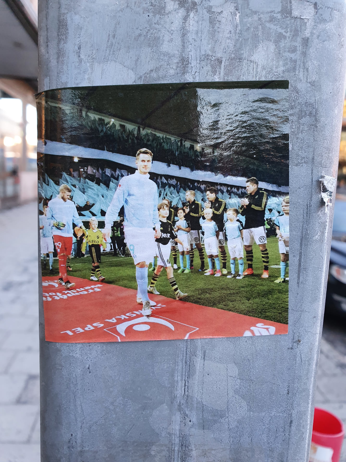 A sticker featuring a photo of a male footballer in a light blue uniform, walking on a red carpet. He is surrounded by a line of young boys, also in light blue uniforms, who appear to be part of a football team.  The background shows a stadium. The sticker has a logo at the bottom that resembles a crescent moon.