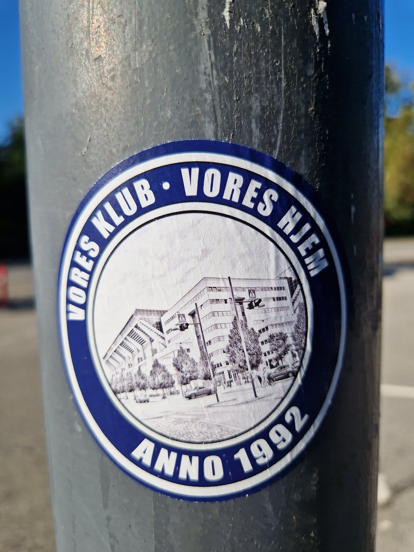 Round blue and white sticker depicting a sketch of a building with the text "Vores Klub Vores Hjem Anno 1992".