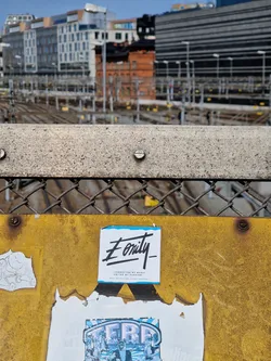 A light blue square sticker with the word 'Eonity' written in a stylized black font.  Beneath the main text, there's smaller text that appears to say 'connection of worlds' and 'driven by passion'. The sticker is affixed to a yellow metal surface, possibly a construction sign, alongside other stickers and posters.