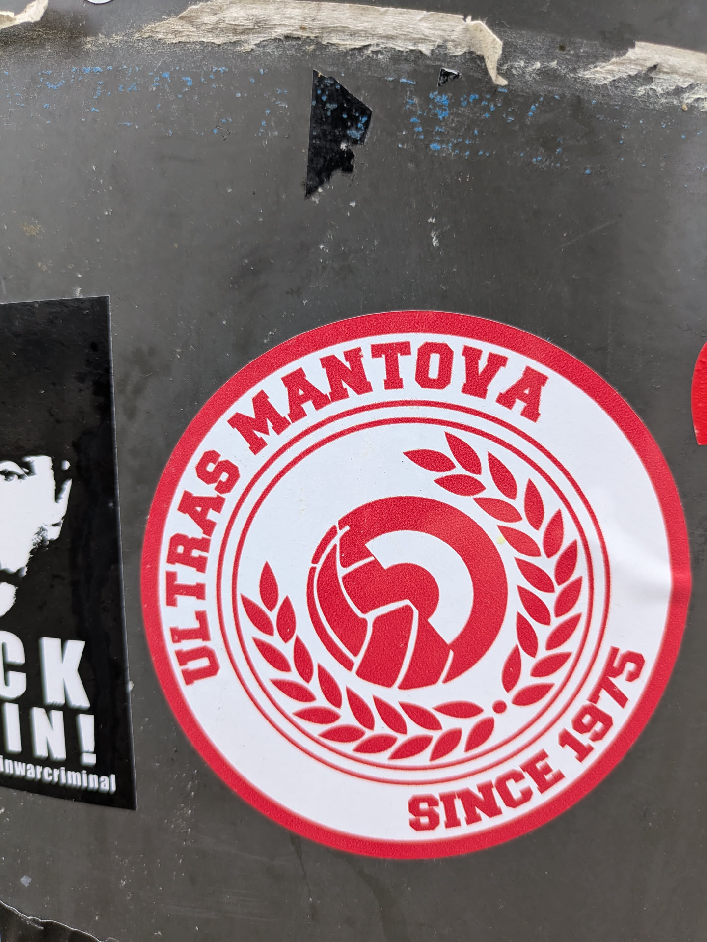 The sticker is round with a red and white color scheme. It has the words "ULTRAS MANTOVA" printed in a circular fashion at the top. At the bottom, it says "SINCE 1975". In the center, there is a stylized depiction of a soccer ball surrounded by laurel leaves.