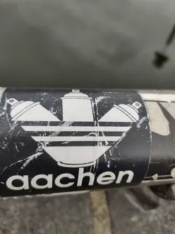 A black and white sticker with a design that combines the Adidas logo with spray paint cans. The word 'aachen' is written at the bottom of the sticker. The sticker shows signs of wear.