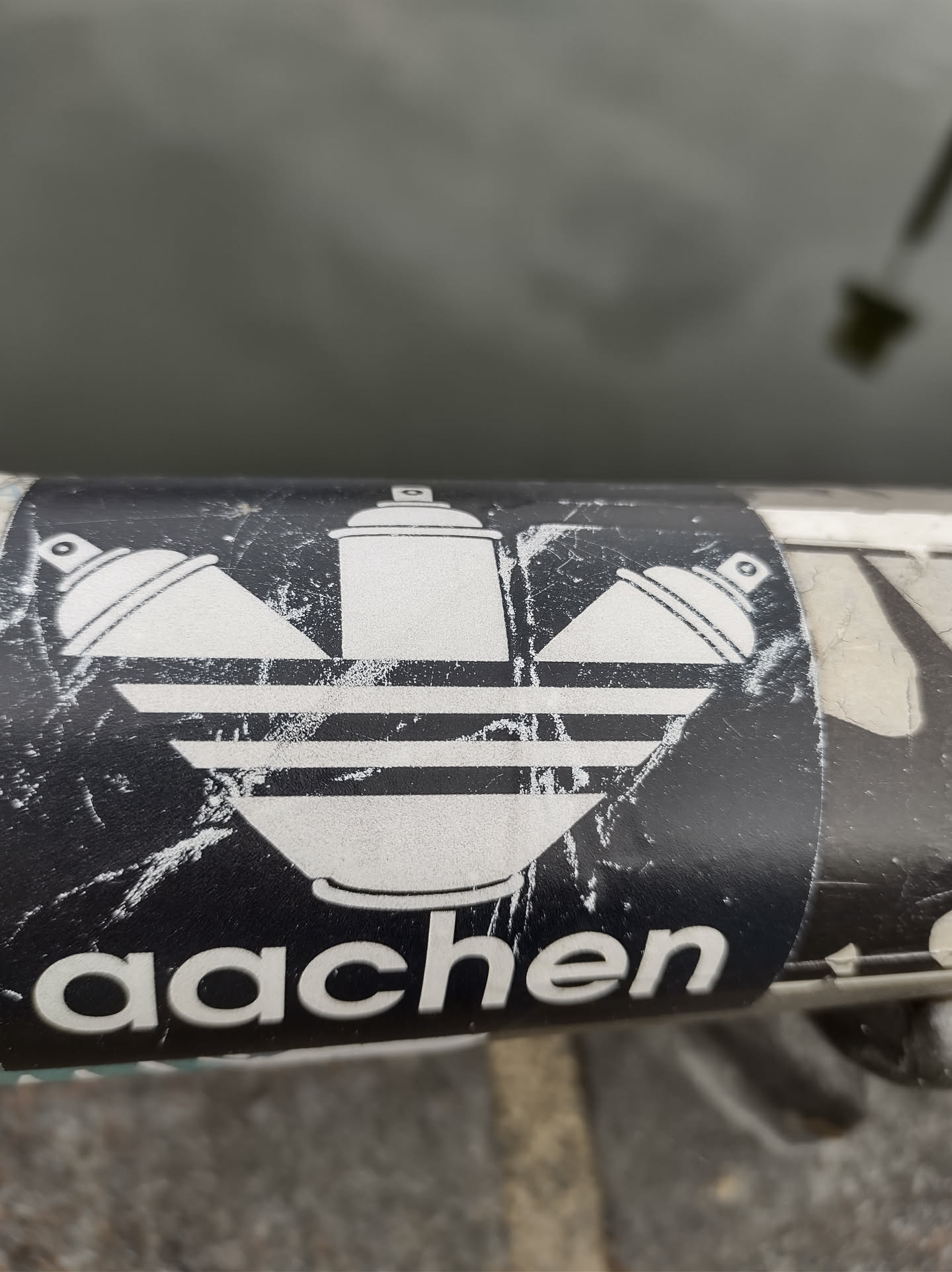 A black and white sticker with a design that combines the Adidas logo with spray paint cans. The word 'aachen' is written at the bottom of the sticker. The sticker shows signs of wear.
