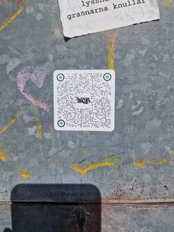 A small, rectangular sticker with a light teal background. The sticker features text written in braille, with a stylized word or logo in a non-braille font in the middle. The top and bottom of the sticker are marked with light teal circles. The sticker is affixed to a worn, textured metallic surface with other graffiti visible nearby. The braille text is densely packed and does not appear to contain a QR code.
