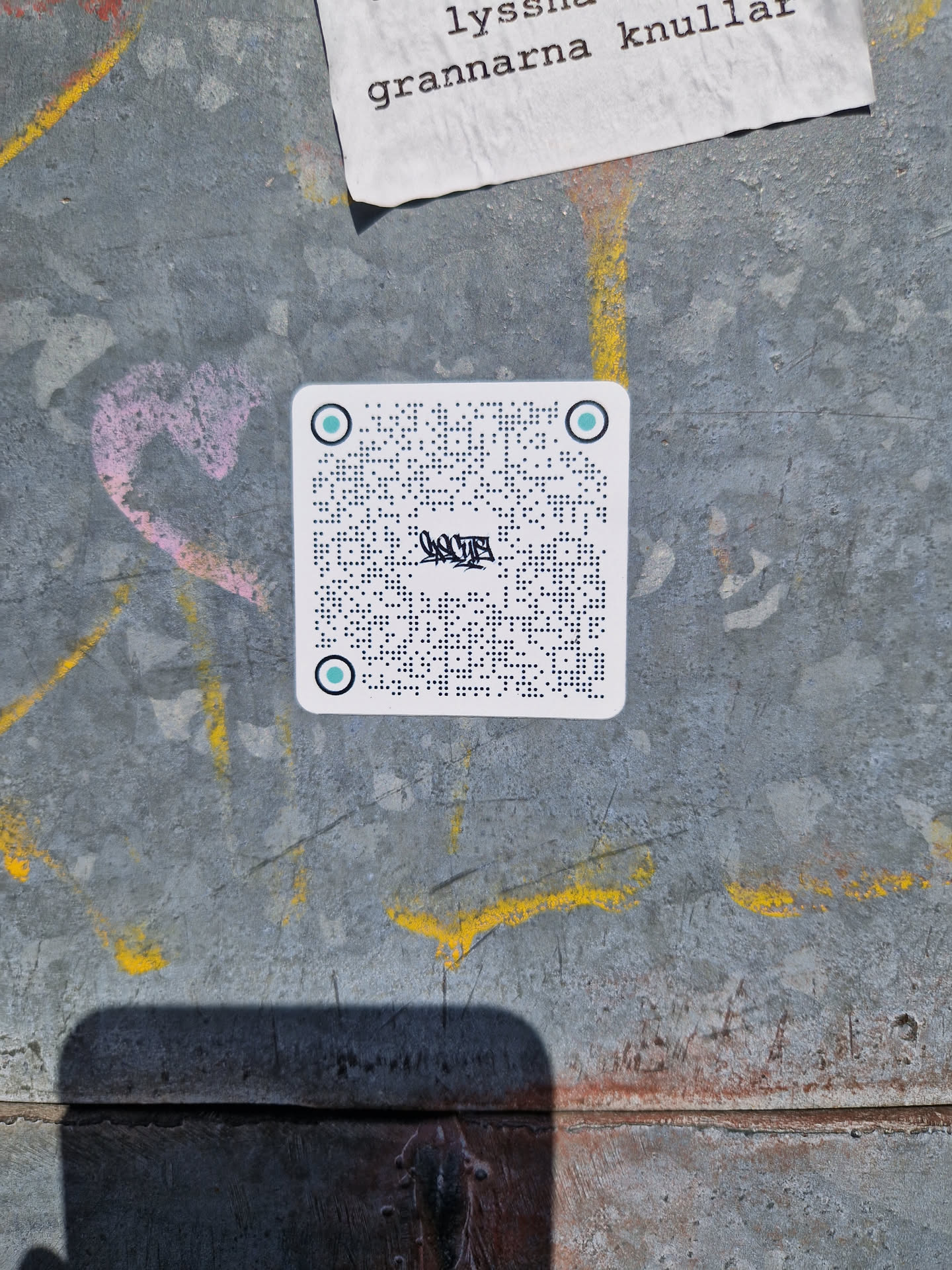 A small, rectangular sticker with a light teal background. The sticker features text written in braille, with a stylized word or logo in a non-braille font in the middle. The top and bottom of the sticker are marked with light teal circles. The sticker is affixed to a worn, textured metallic surface with other graffiti visible nearby. The braille text is densely packed and does not appear to contain a QR code.