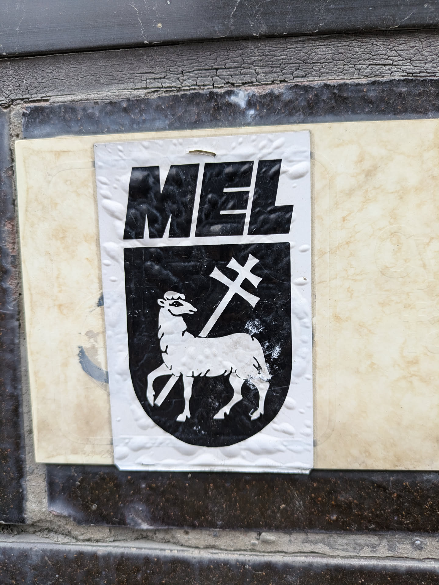 A black and white sticker depicting a heraldic shield with a lamb and a cross. The word "MEL" is written above the shield in bold, uppercase letters.
