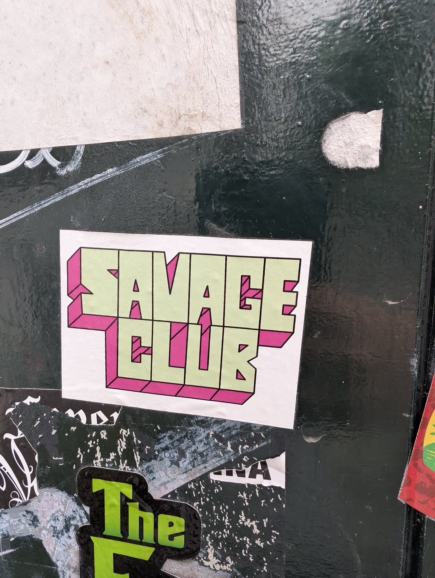 A white square sticker features the words "SAVAGE CLUB" stacked on two lines. The text is rendered in a light green fill with a bold pink outline, giving a three-dimensional, retro vibe. The sticker is pasted on a weathered surface, with other stickers and graffiti visible around it.