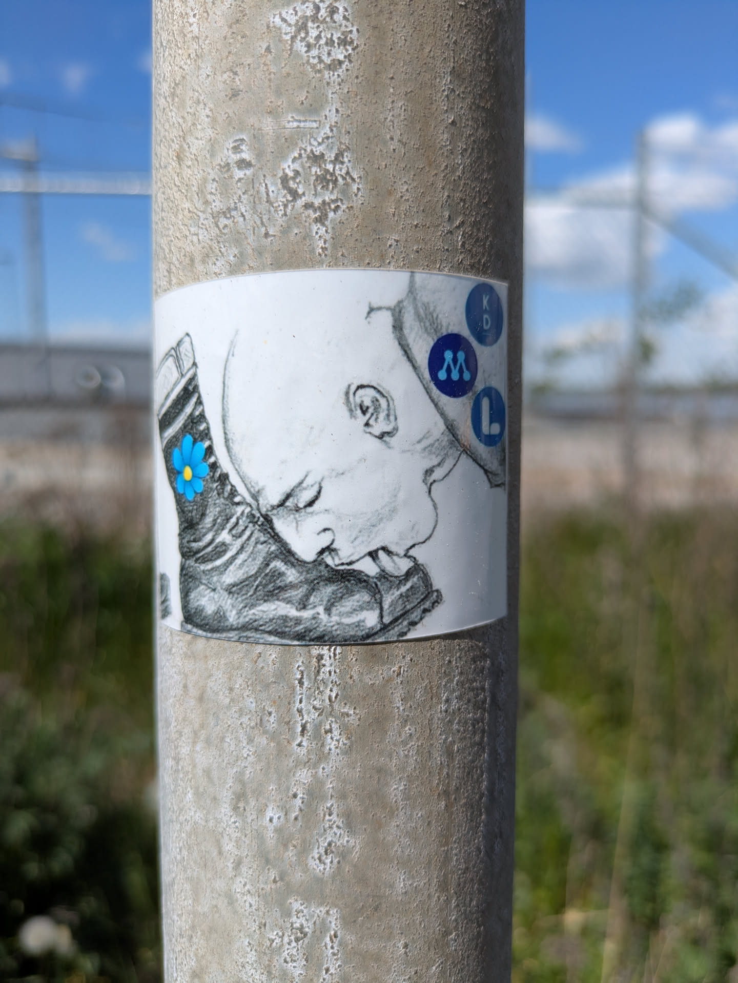 This sticker shows a black and white sketch of a person licking a boot. The boot has a blue flower on it. To the right of the head is a vertical stack of blue circles, each containing a letter: K, D, M, and L.