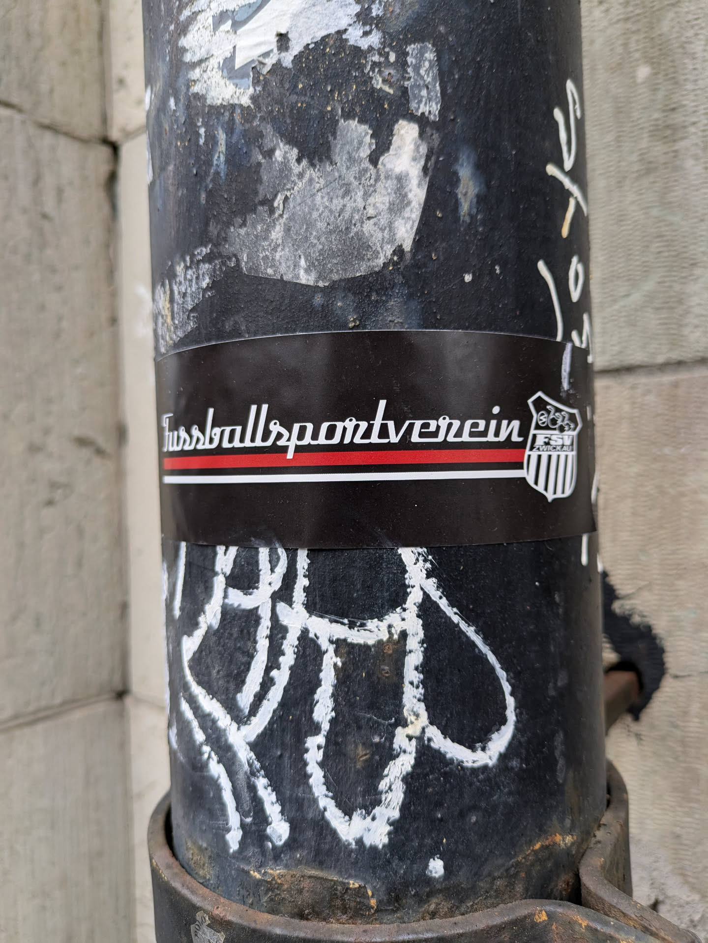 A black and white sticker featuring the text "Fussballsportverein" in a stylized font. A small emblem is visible on the right.  The sticker is affixed to a dark metal post covered in other graffiti and markings.