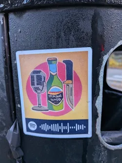 Street sticker A rectangular sticker with rounded corners and a white border affixed to a textured black surface. The upper graphic area has a muted yellowish background featuring a central pink circle. In front of the circle are illustrations of three objects: a vintage silver microphone on a stand, a green beer-style bottle labeled 'KRIMINAL TANGO', and an upright hunting knife with a dark blue or black handle. Below the main graphic is a dark navy horizontal bar displaying the Spotify logo alongside a scannable Spotify waveform code.