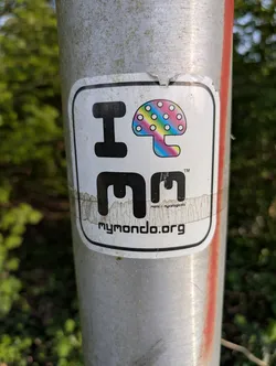 A white sticker with the words "I Mondo" and a rainbow mushroom. The sticker promotes mymondo.org. It is affixed to a metal pole.