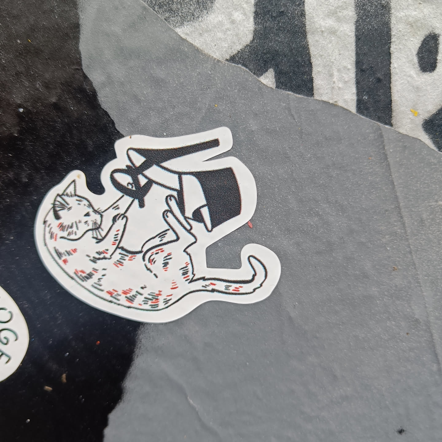 A sticker featuring a cartoon drawing of a cat playfully interacting with a high heel shoe. The cat is depicted in a dynamic pose, suggesting movement and interaction. The style is simple, with black outlines and minimal color accents.