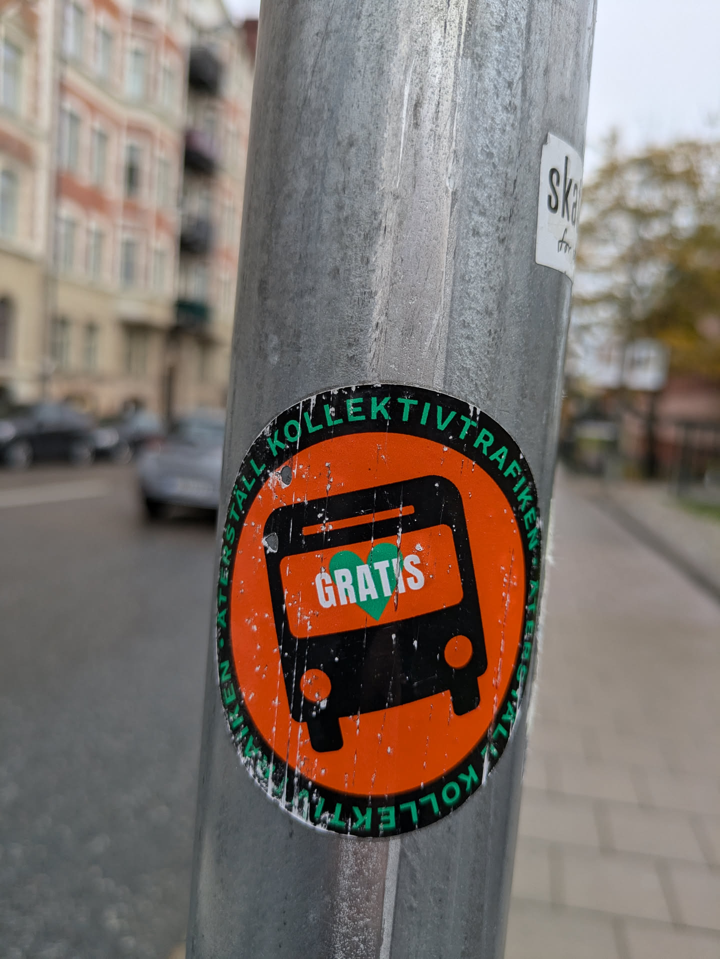 An orange and green round sticker stuck on a metal pole.  The sticker features a simple illustration of a bus with the word "GRATIS" (meaning "free" in German) written on it.  The text "KOLLKEKTIVTRAFIK" is written around the circular design, likely meaning "public transport" in Swedish.
