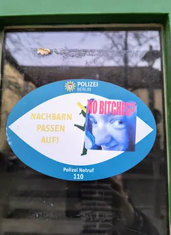 Oval-shaped sticker on a glass door. It has a blue background and contains text in German that translates to "Neighbors, watch out!" There's also a smaller sticker overlaid that reads "NO Bitches?" with a picture of a woman.  The larger sticker also includes the Berlin Police emergency number.