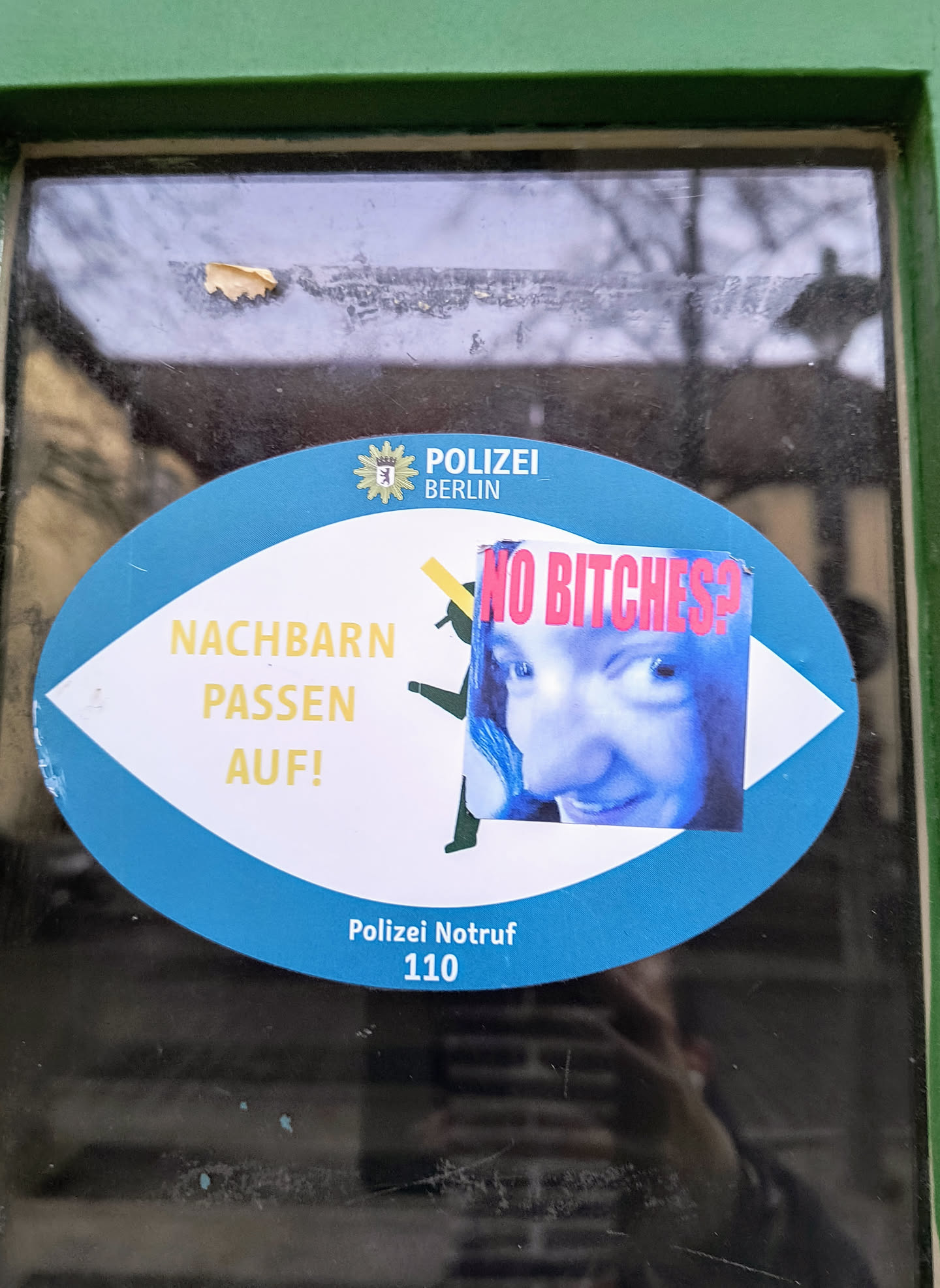 Oval-shaped sticker on a glass door. It has a blue background and contains text in German that translates to "Neighbors, watch out!" There's also a smaller sticker overlaid that reads "NO Bitches?" with a picture of a woman.  The larger sticker also includes the Berlin Police emergency number.
