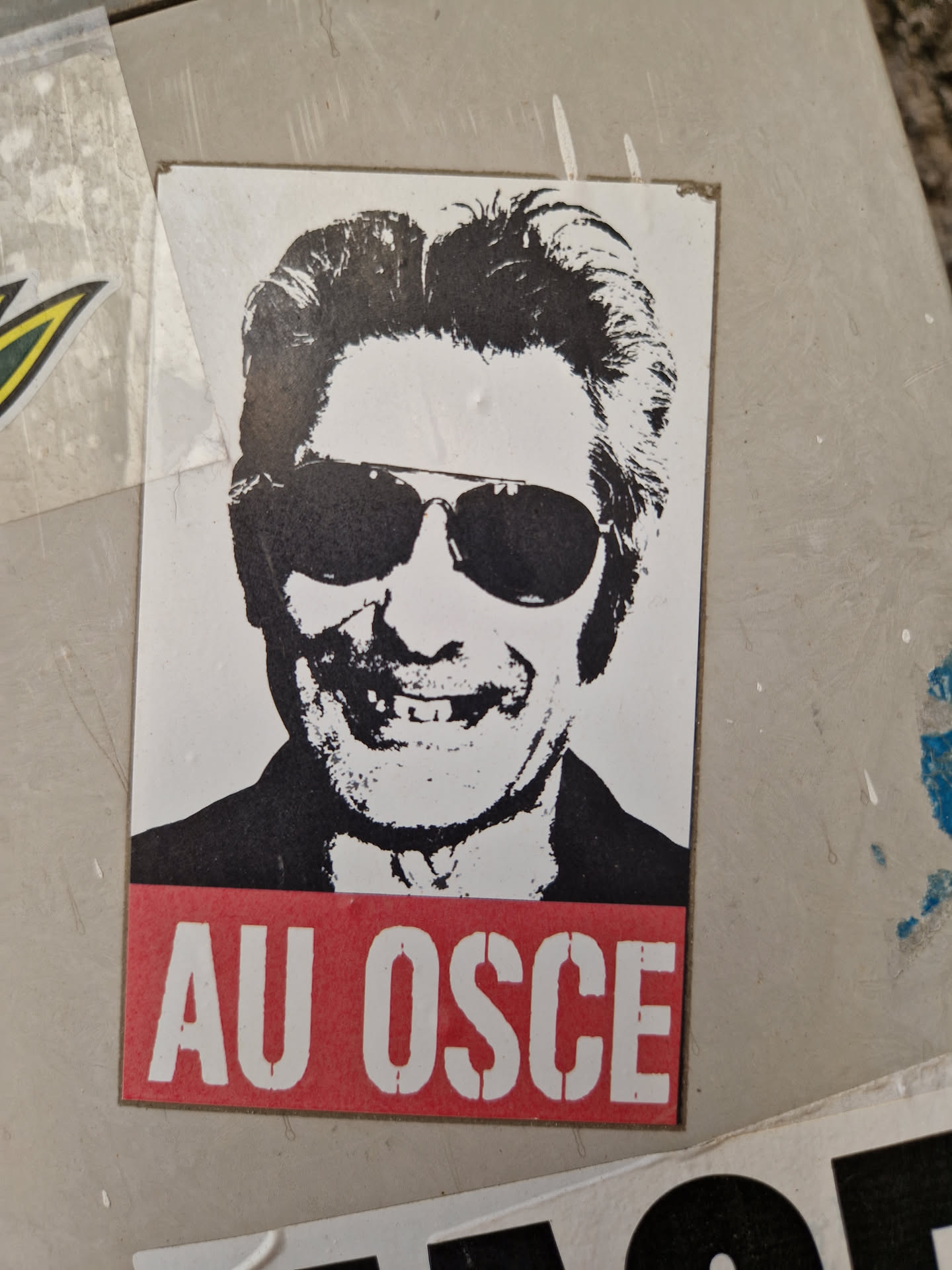 Black and white sticker featuring a stylized portrait of a man wearing sunglasses.  The text 'AU OSCE' is displayed prominently at the bottom in red.