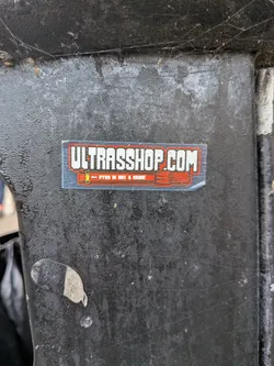 Street sticker A rectangular transparent adhesive sticker stuck onto a weathered black metal surface. The upper section features large, bold white typography outlined in orange that reads 'ULTRASSHOP.COM'. Directly underneath is a stylized orange and red illustration of a handheld flare casing with an arrow and the white text 'PYRO IS NOT A CRIME' running horizontally along its body.