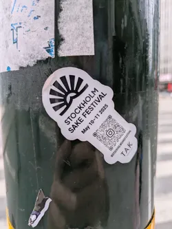 A distinctive black and white die-cut sticker shaped somewhat like a sake tokkuri (flask) with a rising sun motif at its top. The sticker advertises the 'Stockholm Sake Festival' scheduled for 'May 10-11 2025'. It prominently features a QR code with an embedded Instagram logo, alongside the handle '@STOCKHOLMSAKEFESTIVAL'. Below the main information, the text 'TAK' and 'STOCKHOLM | OSLO' are visible. The sticker is affixed to a dark green, possibly metallic, pole, with older, torn paper remnants visible around it.