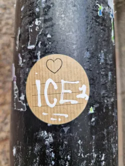 A round brown sticker with the word ICE written on it in white. There is a small heart drawn above the word ICE. The sticker is attached to a dark-colored surface.