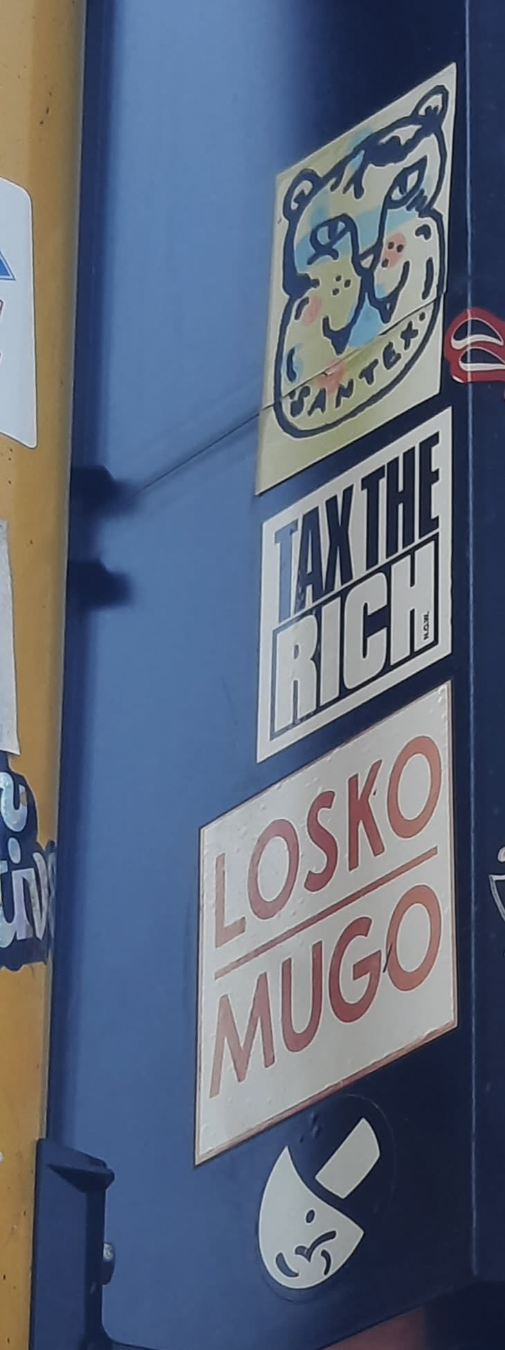 A rectangular sticker with a white background and a thick black border. It features the bold, block text "TAX THE RICH" in white, with the top line "TAX THE" appearing to have a subtle blue tint or reflection, while "RICH" is solid white. To the right of "RICH", there's small, faint text that appears to read "N.C.W.L.". The sticker conveys a clear political message.