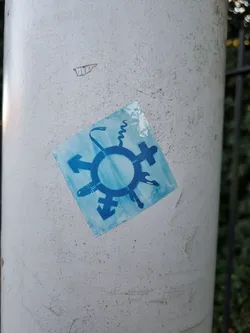 A square blue sticker with a symbol representing different gender identities is affixed to a light gray pole. The symbol is composed of a circle surrounded by arrows and other gender signs.
