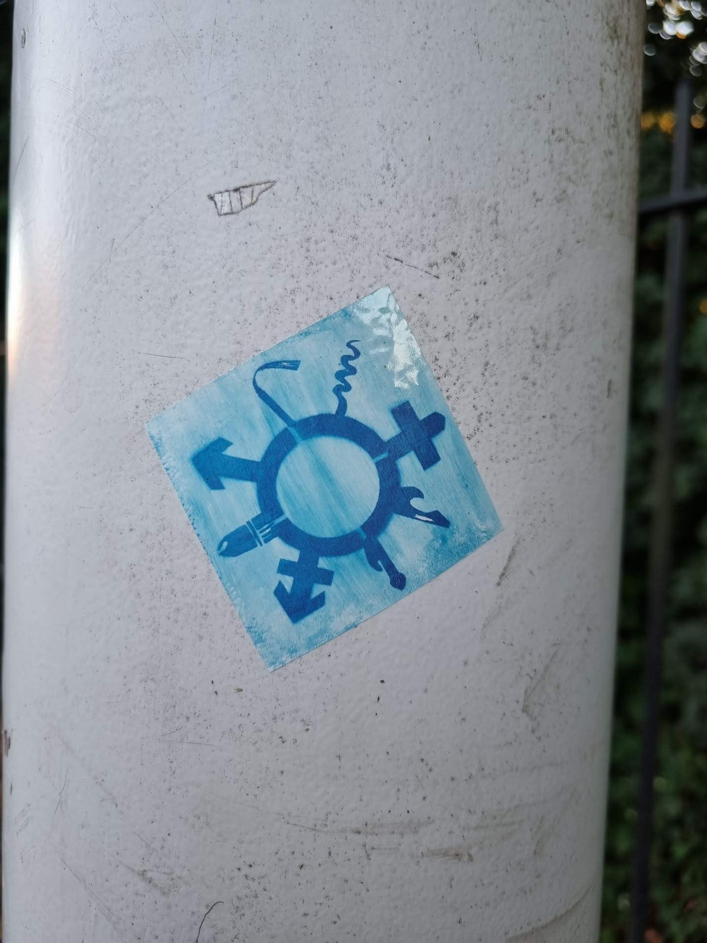 A square blue sticker with a symbol representing different gender identities is affixed to a light gray pole. The symbol is composed of a circle surrounded by arrows and other gender signs.