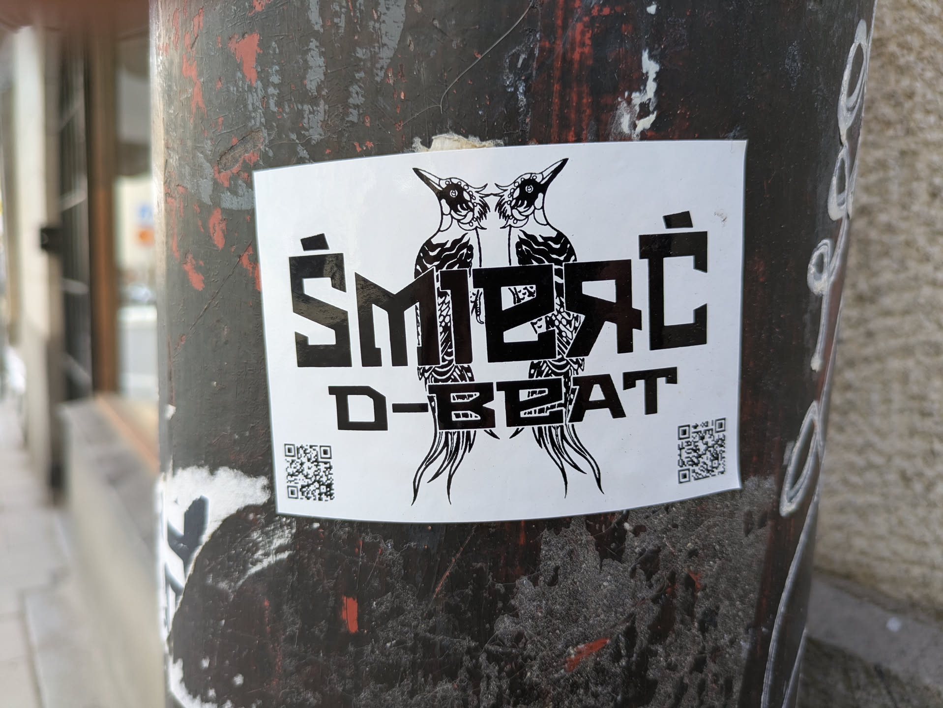 The sticker features two woodpeckers facing each other above the text "ŚMIERĆ D-BEAT". The text is in a bold, blocky font. There are QR codes on the bottom left and right corners of the sticker.