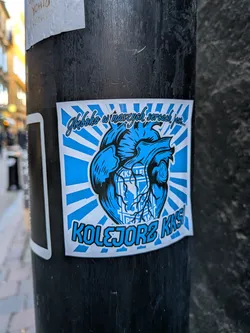 A square sticker featuring an illustrated anatomical heart in blue and black line art against a radiating blue and white sunburst background. The emblem of the Polish football club KKS Lech Poznań is subtly integrated across the front of the heart. Above the heart, cursive script text reads 'Głęboko w naszych sercach jest...', and beneath it, bold block lettering with black borders reads 'KOLEJORZ KKS!'. The sticker is adhered to a black outdoor pole.