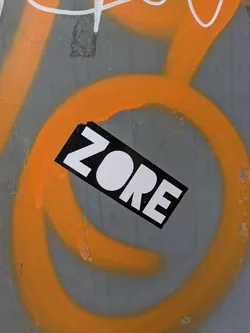 A rectangular sticker with the text "ZORE" in white letters on a black background is affixed to a gray surface covered in orange graffiti.