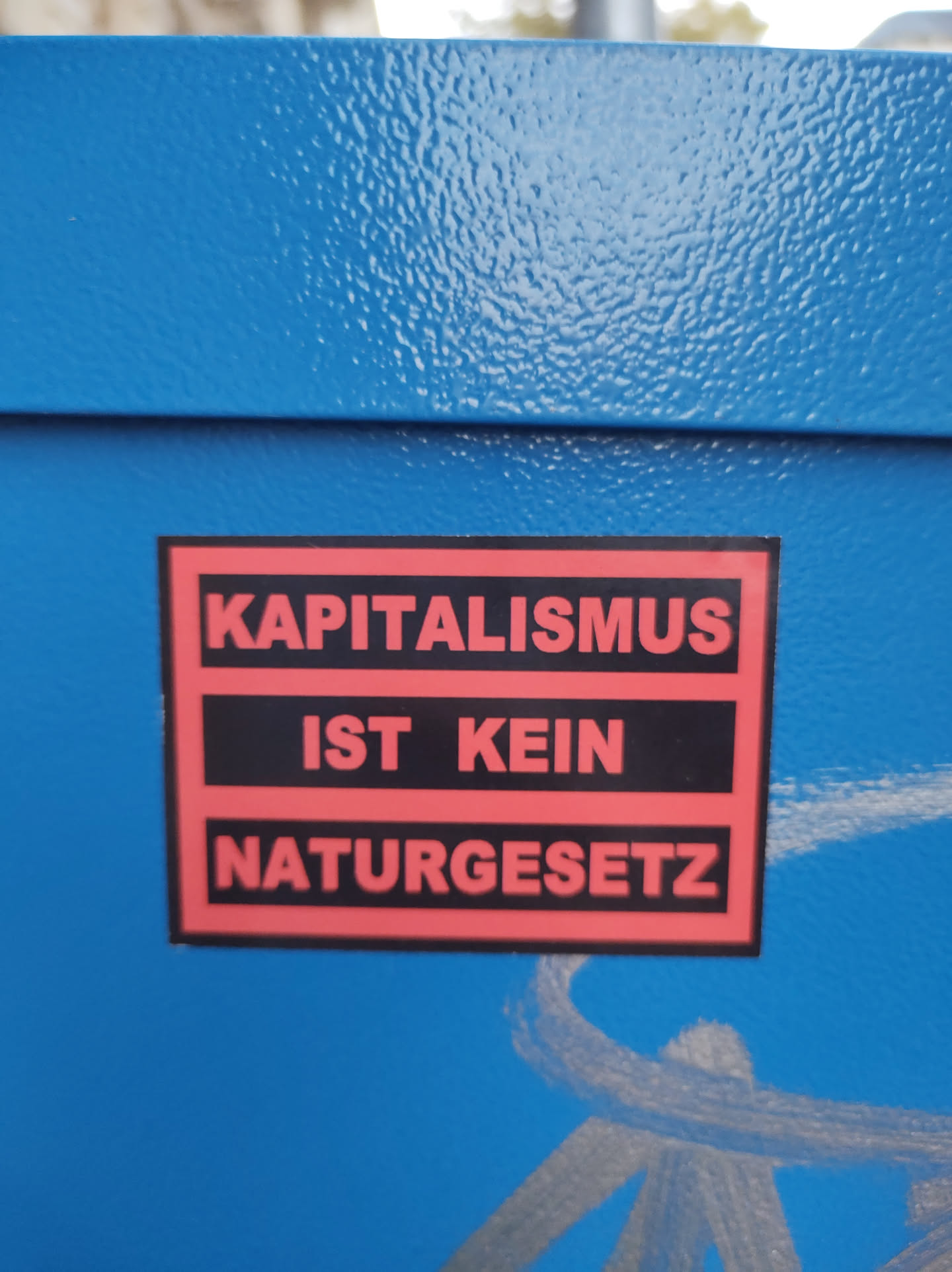 A sticker with the text "Kapitalismus ist kein Naturgesetz" (Capitalism is not a law of nature) in German.  The sticker has a red and black color scheme and is affixed to a blue surface.