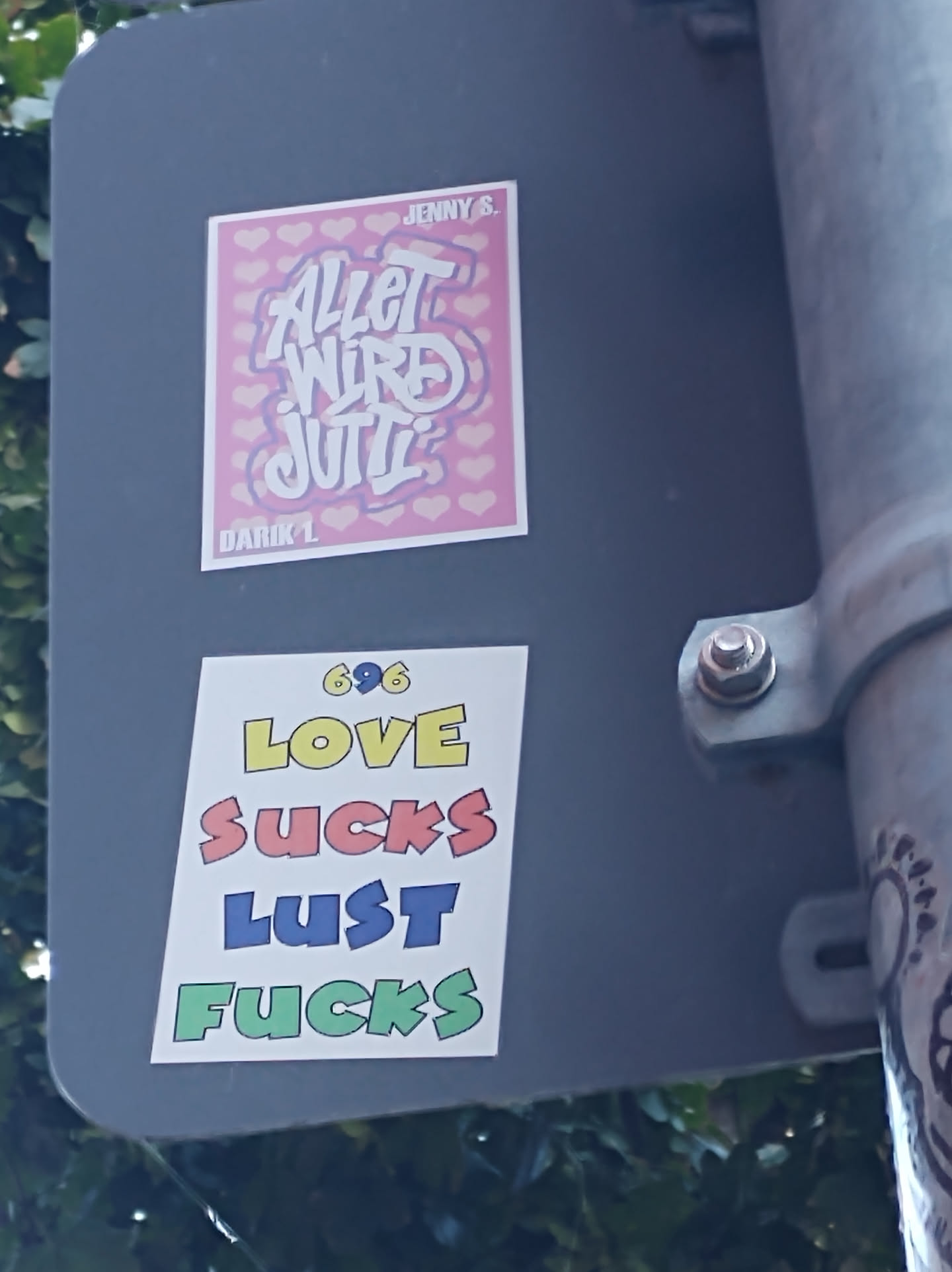 A sticker with the text 696 LOVE SUCKS LUST FUCKS in colorful letters. The sticker is attached to a pole.  There is another sticker above it with text in a different style and language.