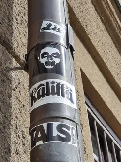 A close-up shot of several stickers affixed to a downspout. The most prominent sticker is a black and white illustration of a face with exaggerated features. Below it is a sticker with the word "kaliffa" in a stylized font. Another sticker at the bottom says "TALSA". The stickers have a somewhat gritty, urban aesthetic. 