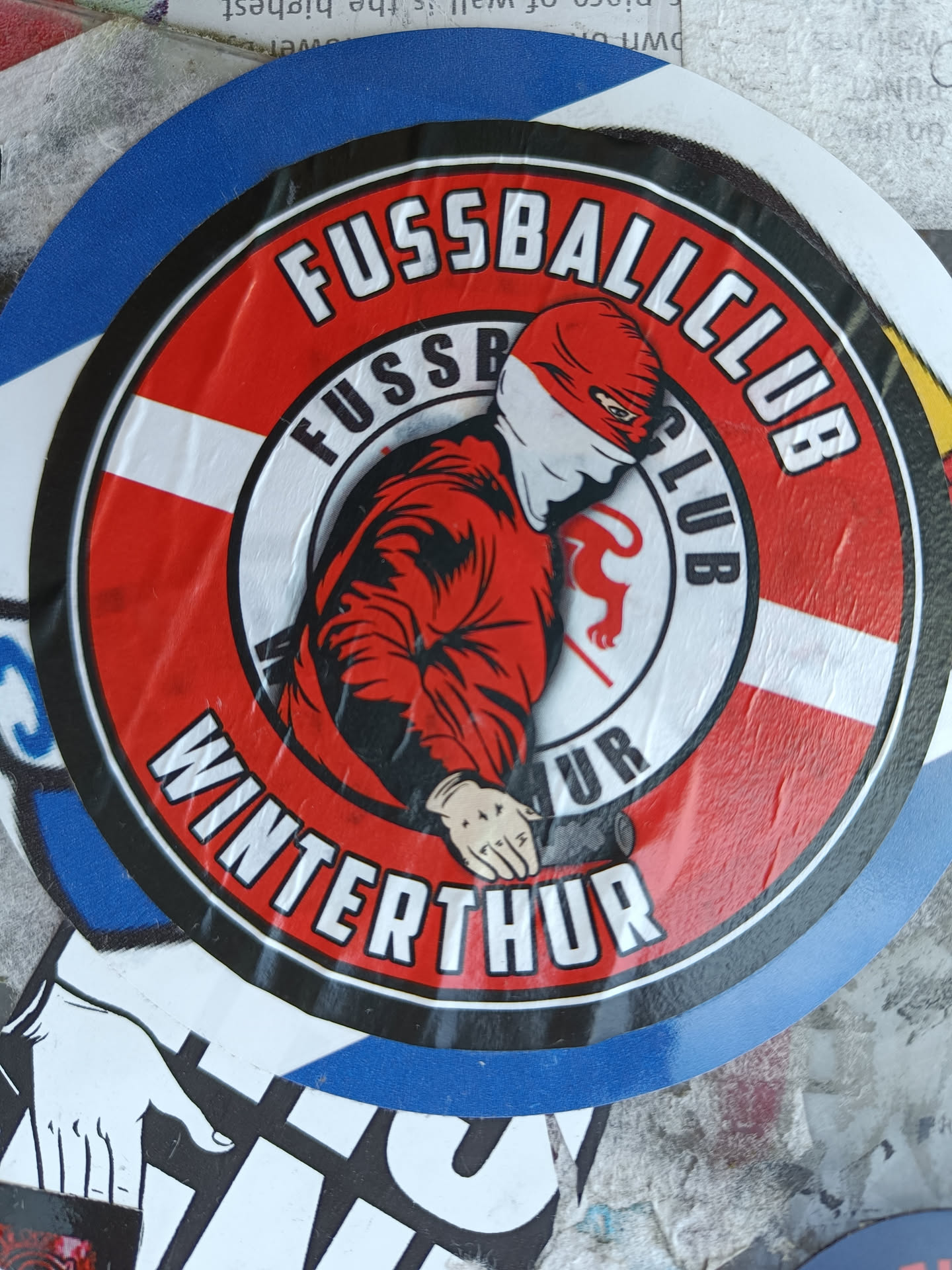 Round sticker featuring a masked figure in a red jacket, the text "Fussballclub Winterthur" and the club's logo. The sticker's design is reminiscent of hooligan or ultras subculture.
