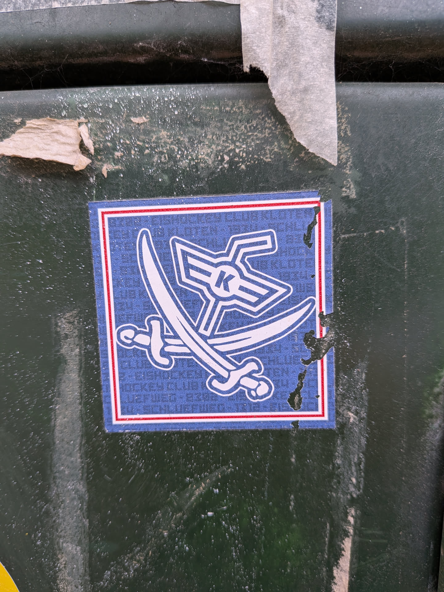 The sticker is square with a blue background and a white border, outlined by a thin red line. In the center, two white crossed swords are depicted, with their hilts pointing downwards. Above the swords is a stylized white emblem that resembles wings or a crest, with a prominent white 'K' in the center. The blue background has a subtle pattern of repeating text, which appears to be 'HOCKEY CLUB' in white, though it is difficult to read clearly. The sticker shows some signs of wear and tear, with a small section of the right edge appearing to be peeling or damaged.