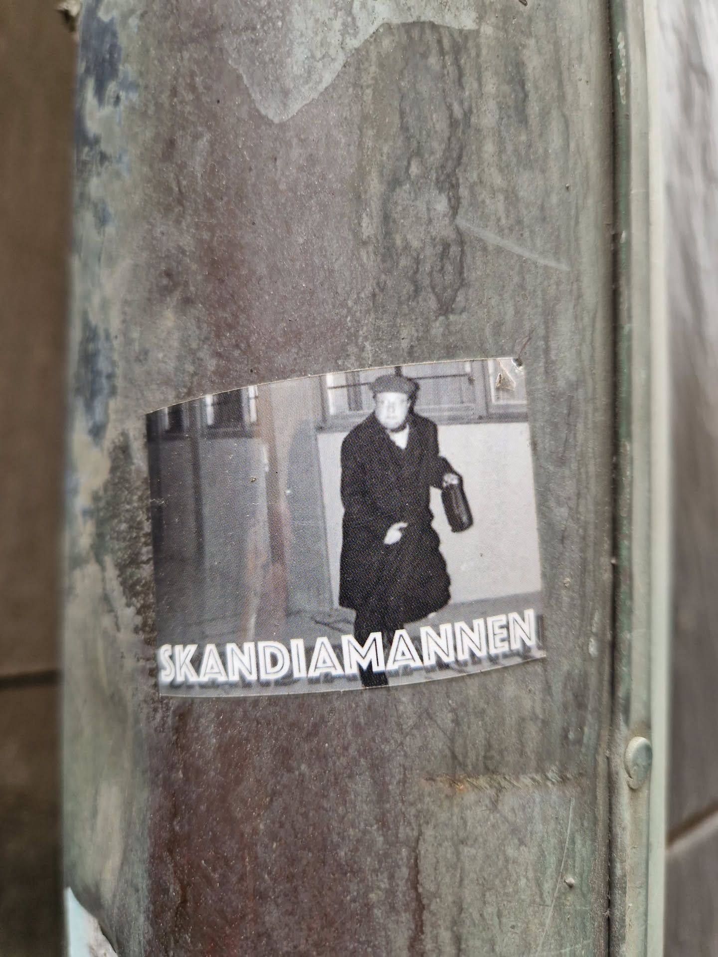 Black and white photo of a man in a coat walking down a street. The sticker has the text "Skandia mannen" at the bottom.