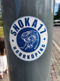 A blue and white circular sticker featuring the text 'Snok A17 Norrköping' and a graphic illustration of what appears to be a snake or serpent within a wave-like design. The sticker is affixed to a grey pole outdoors.