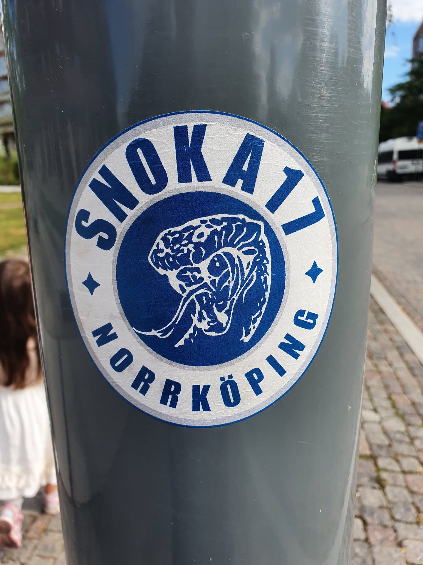 A blue and white circular sticker featuring the text 'Snok A17 Norrköping' and a graphic illustration of what appears to be a snake or serpent within a wave-like design. The sticker is affixed to a grey pole outdoors.