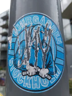 A worn-out blue and white circular sticker featuring a masked figure holding two cylindrical objects. The text "Djurgården Stockholm" is visible around the figure.