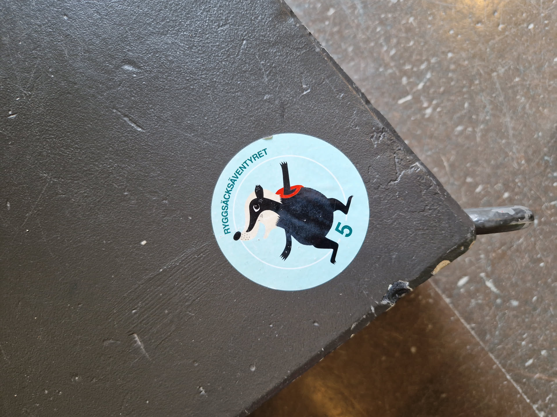A round sticker featuring a cartoon badger in a red scarf, the number 5, and the text "Ryggsacksäventyret" which translates to "Backpack Adventure" in English. The sticker is applied to a dark gray surface.