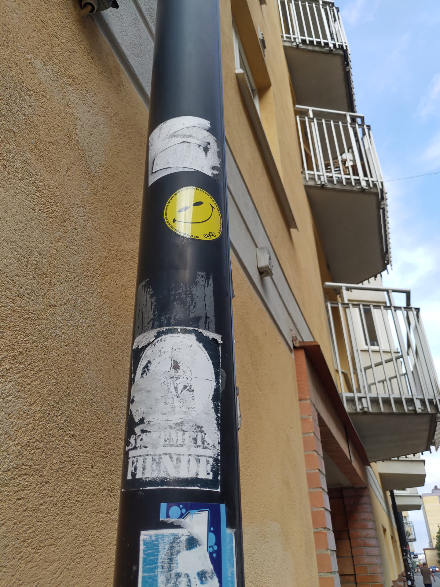 A yellow smiley face sticker with a small detail inside the smile. It is placed on a dark grey pole, amongst other stickers.