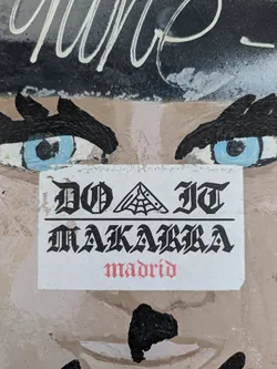 The sticker features the text 'DO IT MAKARRA madrid' in a gothic style font. The word 'DO IT' has a spider web above it, and 'madrid' is written in red. The sticker is peeling and placed on a wall with an unfinished painting of a face around it.