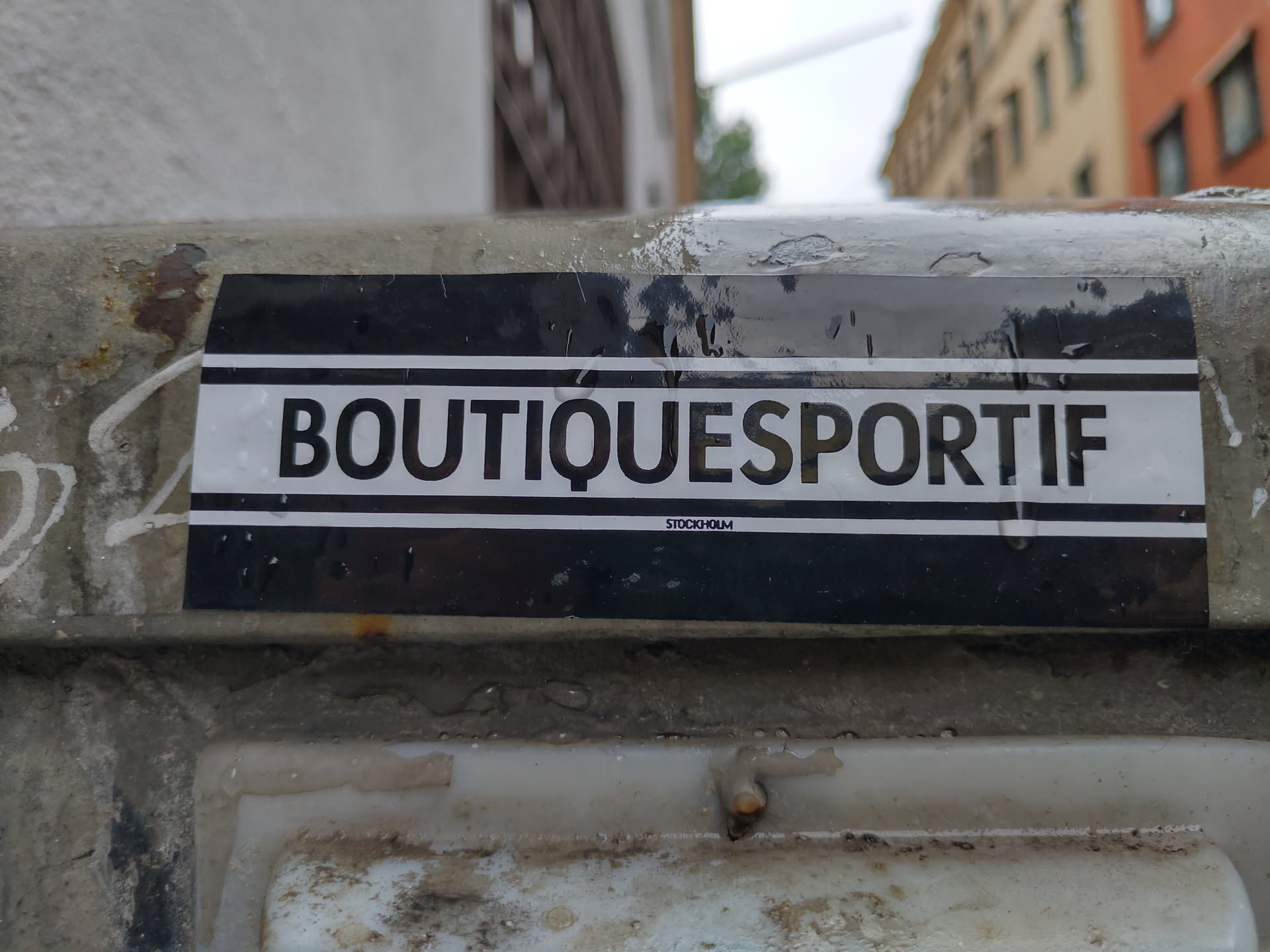 A black and white sticker with the text "BOUTIQUESPORTIF" and "STOCKHOLM" in smaller text underneath. The sticker is attached to a weathered surface.