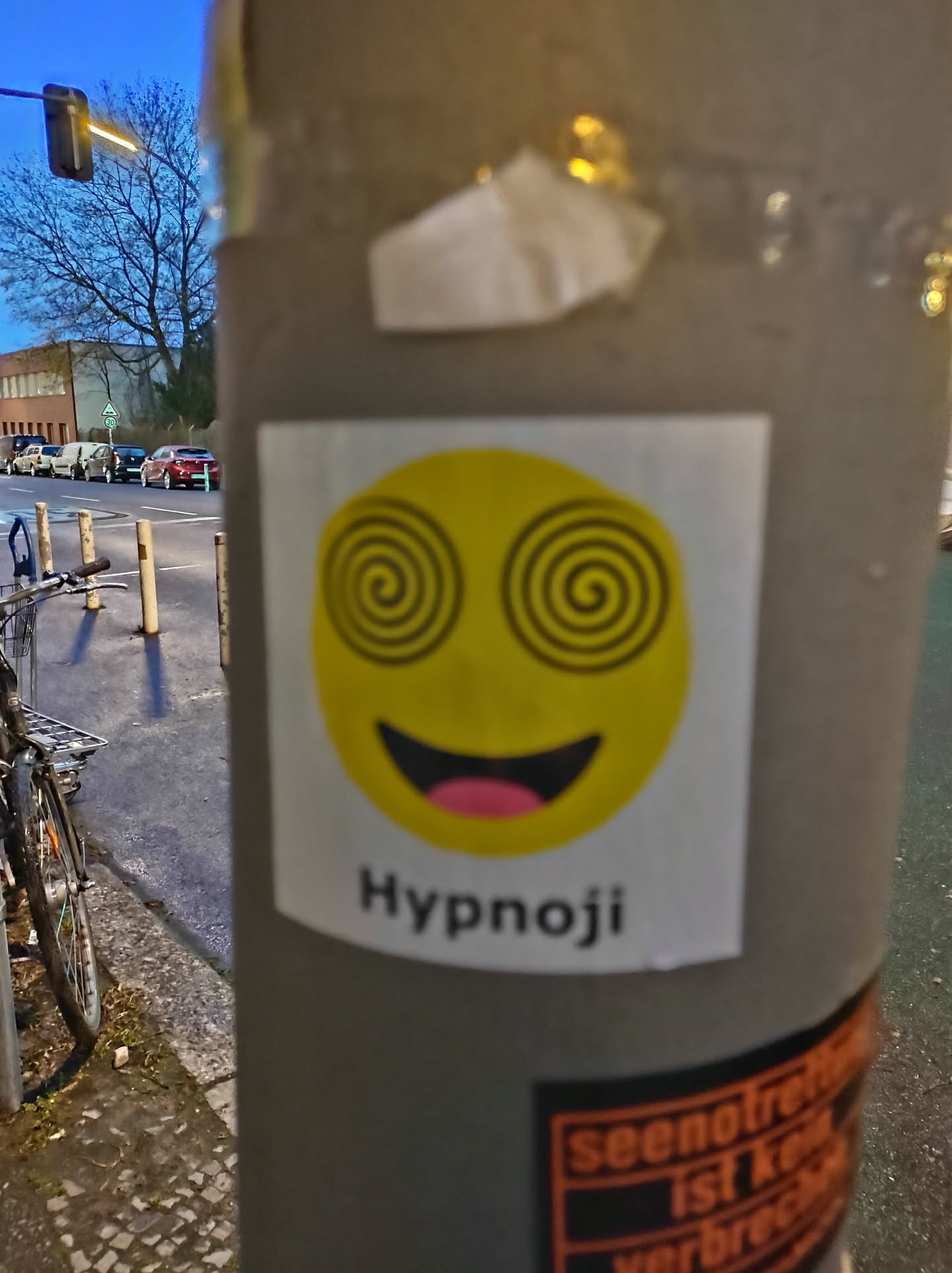 A yellow emoticon sticker with hypnotizing spirals for eyes and a wide open smile. The text 'Hypnoji' is written below the emoticon. The sticker is affixed to a grey pole on a city street.