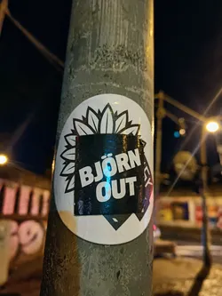 A black and white sticker with the text "BJORN OUT" and a floral design. The sticker is affixed to a weathered gray pole at night.