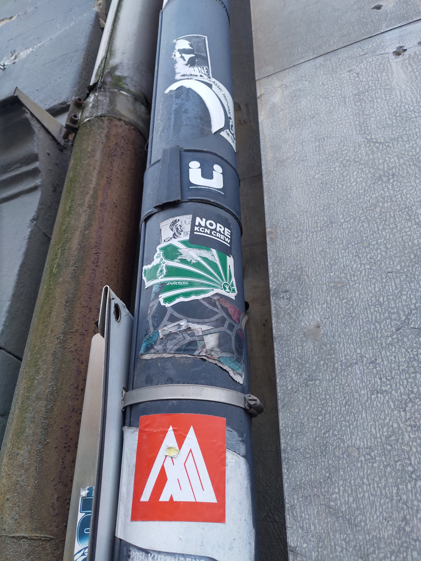A square-shaped sticker with an orange background features a stylized abstract geometric design resembling three interconnected mountains or peaks. The logo is predominantly white or light-colored, creating a striking contrast against the orange backdrop. The sticker appears to be cleanly applied to a gray metal post, among other stickers and urban elements, suggesting an outdoor setting. The logo's style indicates a modern or contemporary design aesthetic, possibly associated with a brand or organization.