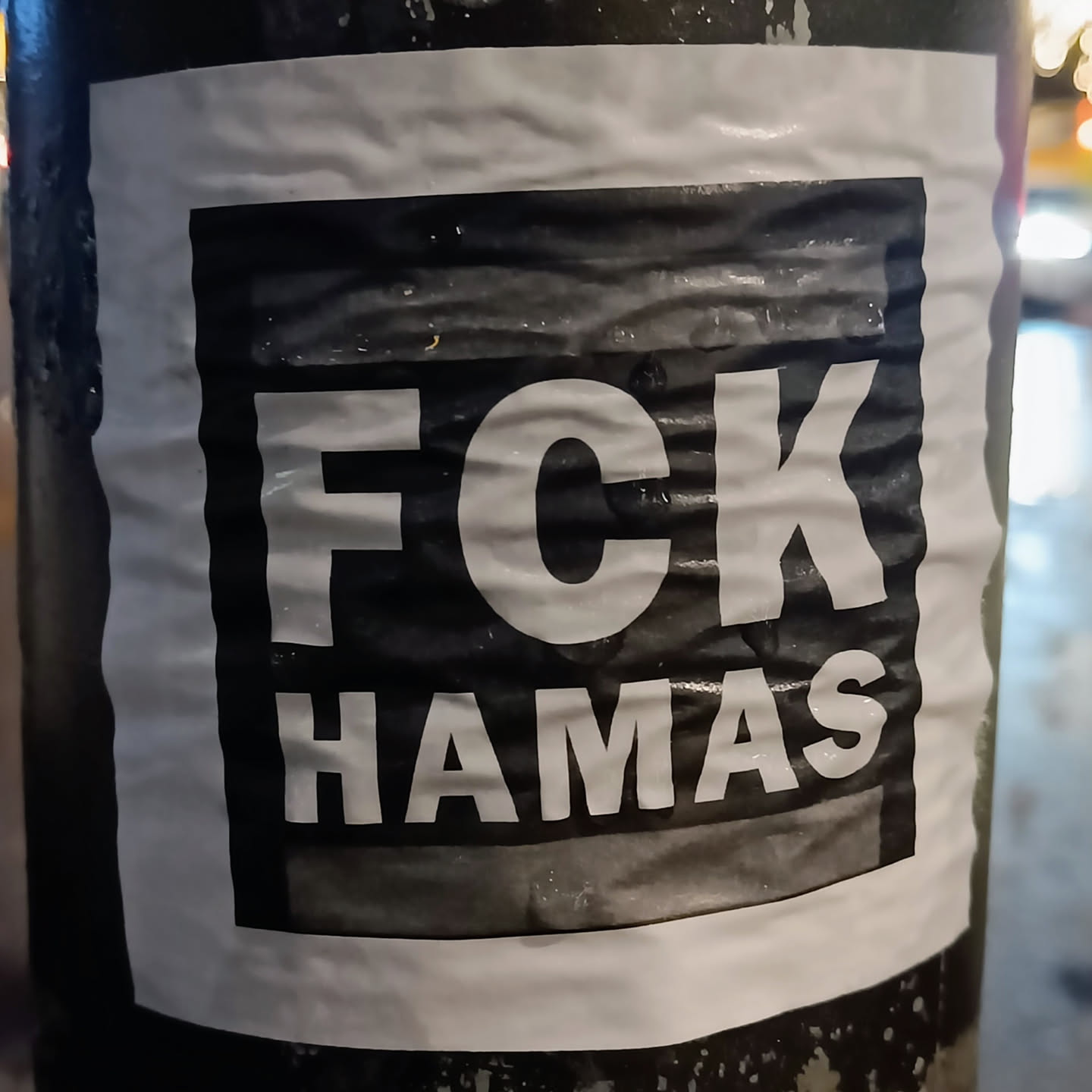 A black and white sticker with the text "FCK HAMAS" printed on it. The sticker is affixed to a dark-colored pole. The sticker appears slightly wrinkled.